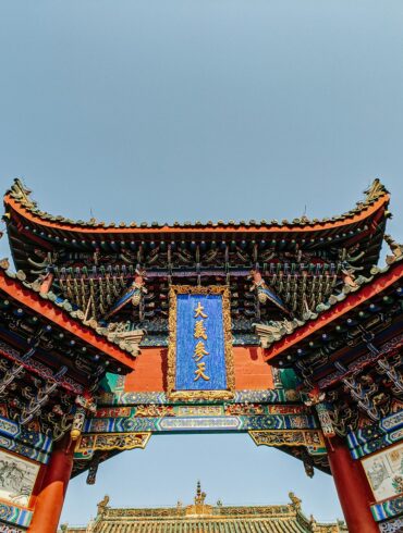 Low-angle view of an ornate traditional Chinese temple gate with curved tiled roofs, red pillars, intricate carvings, and a blue plaque against a clear sky.