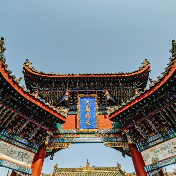 Low-angle view of an ornate traditional Chinese temple gate with curved tiled roofs, red pillars, intricate carvings, and a blue plaque against a clear sky.