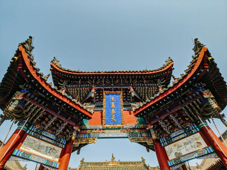 Low-angle view of an ornate traditional Chinese temple gate with curved tiled roofs, red pillars, intricate carvings, and a blue plaque against a clear sky.