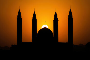 Silhouette of mosque dome and minarets against an orange sunset sky during Ramadan.