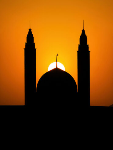 Silhouette of mosque dome and minarets against an orange sunset sky during Ramadan.