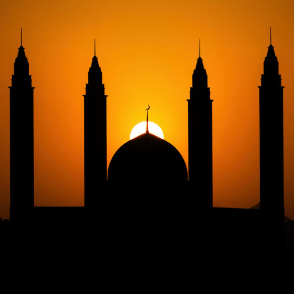 Silhouette of mosque dome and minarets against an orange sunset sky during Ramadan.