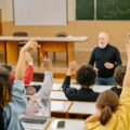 Students raising their hands in a university lecture hall while an older male instructor engages with the class, with laptops open on the desks.