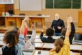 Students raising their hands in a university lecture hall while an older male instructor engages with the class, with laptops open on the desks.