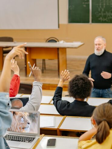 Students raising their hands in a university lecture hall while an older male instructor engages with the class, with laptops open on the desks.
