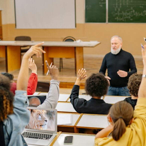 Students raising their hands in a university lecture hall while an older male instructor engages with the class, with laptops open on the desks.