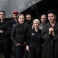 Members of indie band Belle and Sebastian standing together in dark clothing against a modern glass building, looking toward the camera.