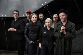 Members of indie band Belle and Sebastian standing together in dark clothing against a modern glass building, looking toward the camera.