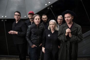 Members of indie band Belle and Sebastian standing together in dark clothing against a modern glass building, looking toward the camera.