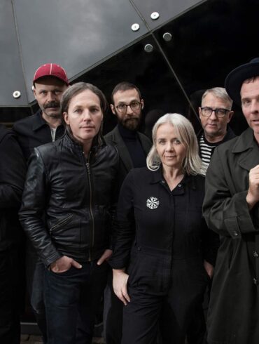 Members of indie band Belle and Sebastian standing together in dark clothing against a modern glass building, looking toward the camera.