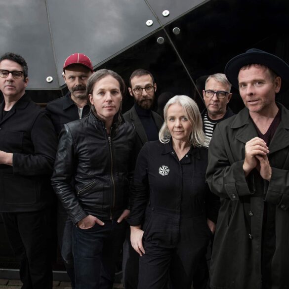 Members of indie band Belle and Sebastian standing together in dark clothing against a modern glass building, looking toward the camera.