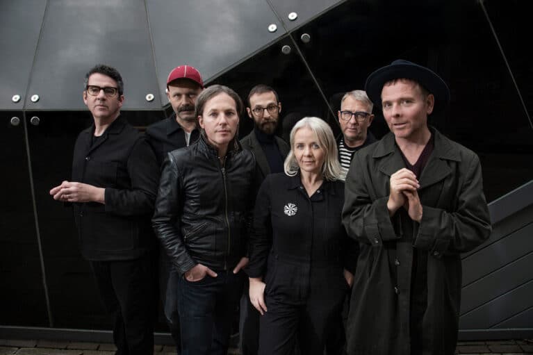 Members of indie band Belle and Sebastian standing together in dark clothing against a modern glass building, looking toward the camera.