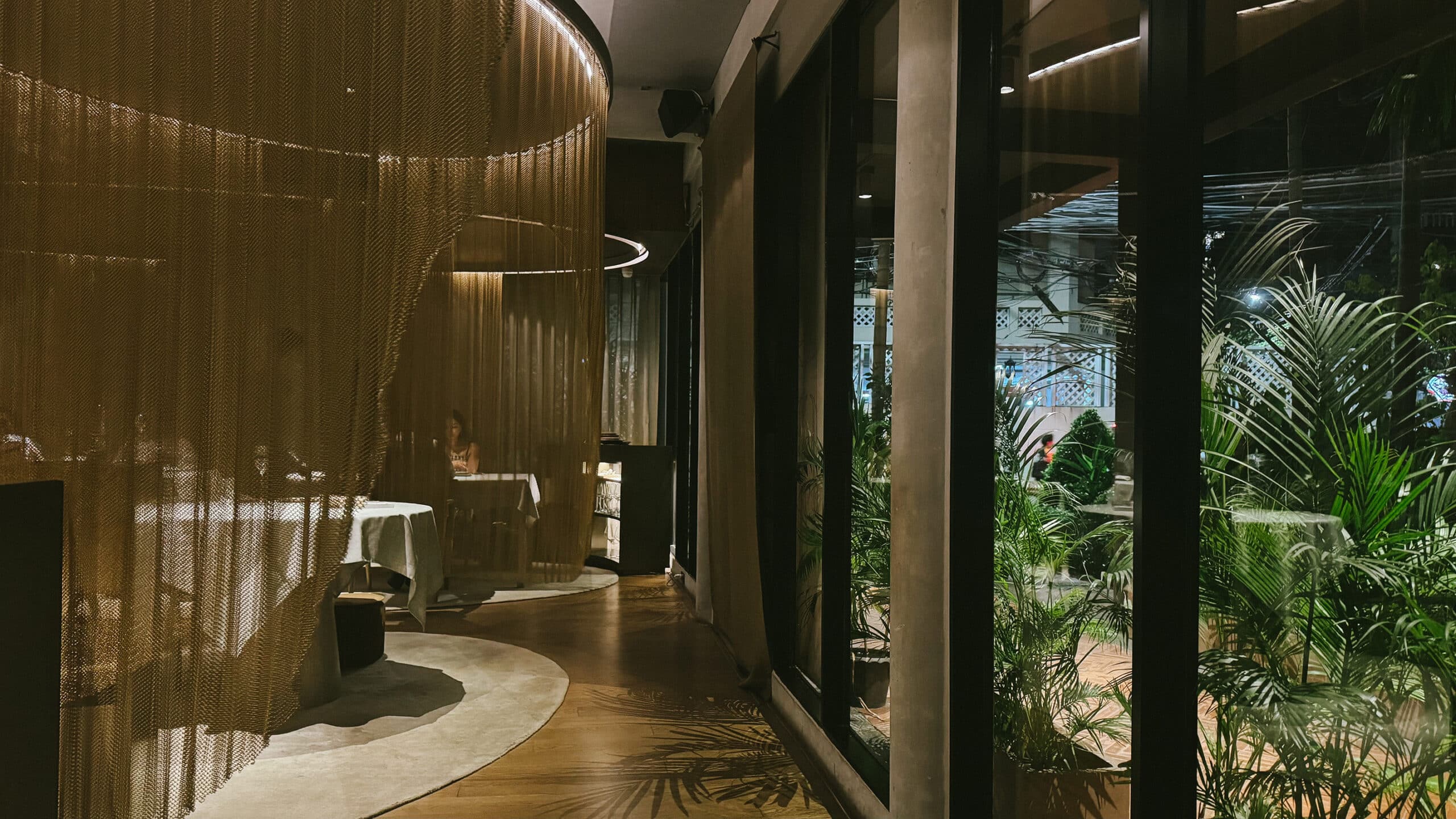 Curved metal chain curtains forming semi-private dining cocoons inside Gaa Bangkok, with warm lighting and views of the garden terrace.
