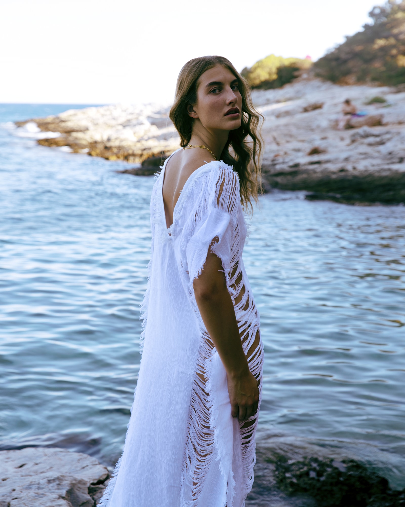 Model wearing a flowing white resortwear dress by Palmier Ile on a rocky seaside coast.