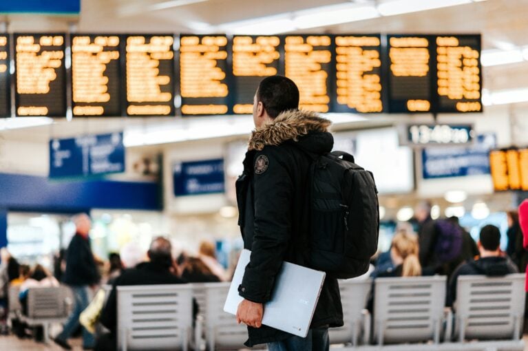 A male traveller holding a laptop looks at a flight information board at an airport terminal.