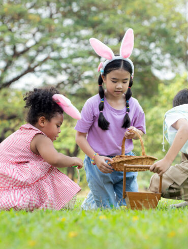 Children wearing bunny ears searching for Easter eggs in a park, holding baskets during an outdoor Easter egg hunt.