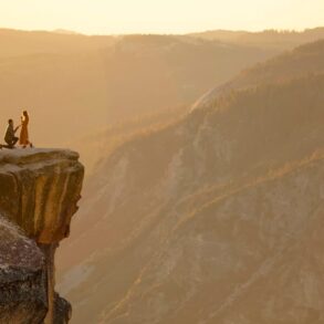Man proposing on one knee to a woman on a cliff edge overlooking a mountain valley at sunset.