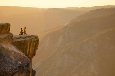 Man proposing on one knee to a woman on a cliff edge overlooking a mountain valley at sunset.