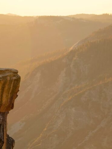 Man proposing on one knee to a woman on a cliff edge overlooking a mountain valley at sunset.