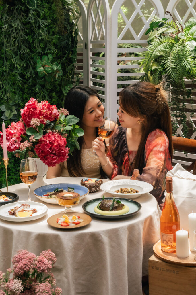 Two women sharing a meal at a flower-adorned table with multiple dishes and rosé wine in a garden-like setting