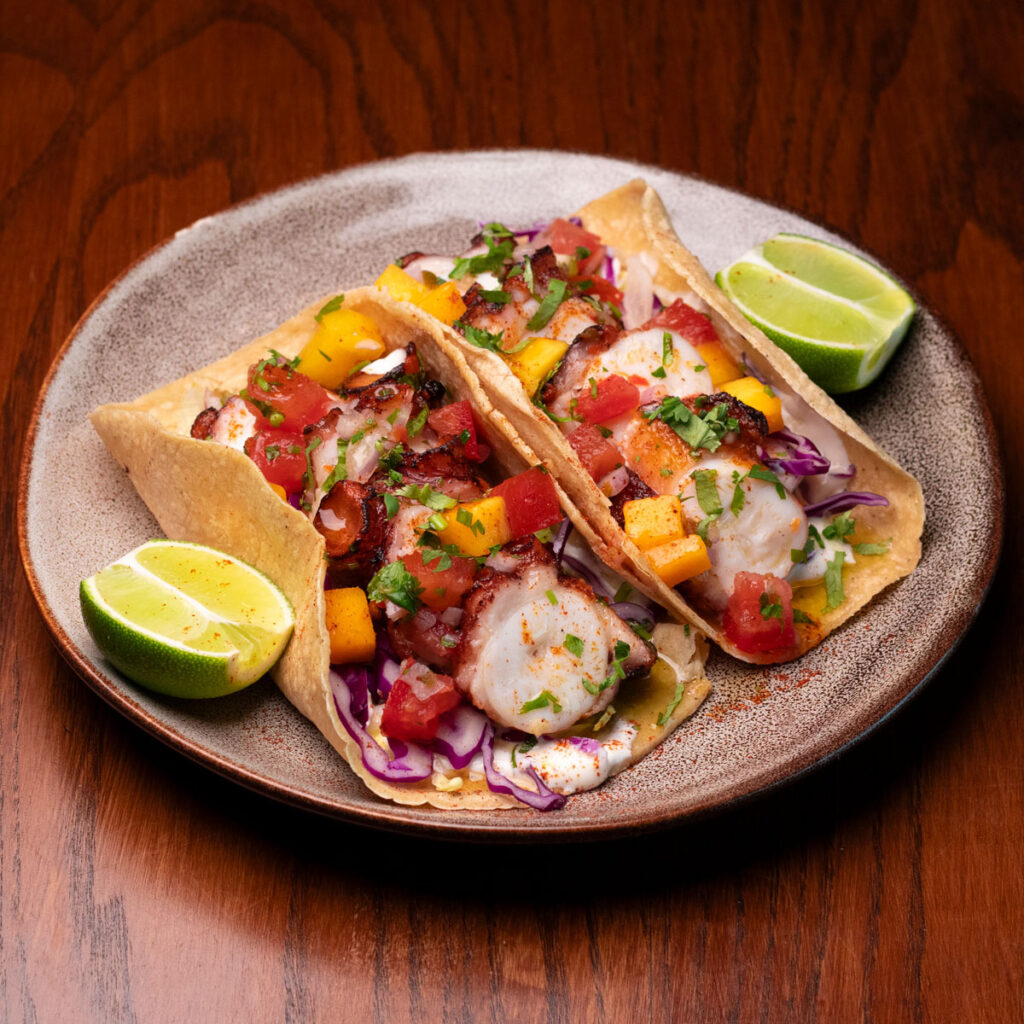 Two grilled octopus tacos on a ceramic plate, topped with mango salsa, pico de gallo, red cabbage and fresh coriander, served with lime wedges at Chimi's Especial, Singapore.