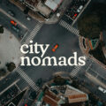 A top view photo of a road unction with motorcycles and cars waiting in traffic lights and an orange car crossing it in Ho Chi Minh Vietnam, with City Nomads logo superimposed.