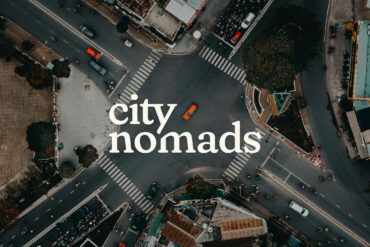 A top view photo of a road unction with motorcycles and cars waiting in traffic lights and an orange car crossing it in Ho Chi Minh Vietnam, with City Nomads logo superimposed.