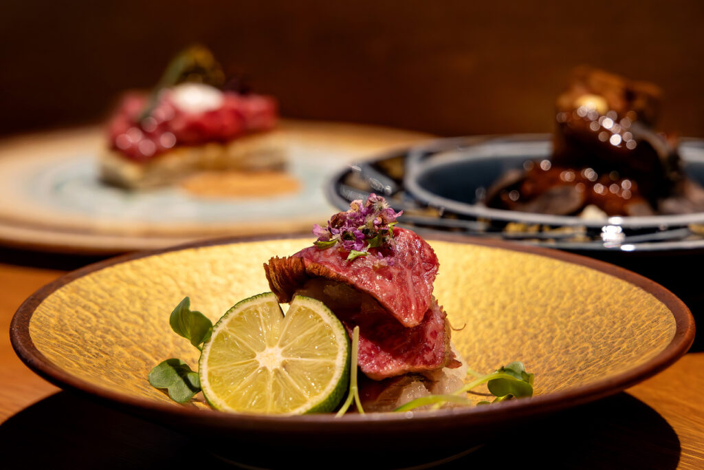 A piece of straw-seared Akune Gold A5 Wagyu tataki topped with edible flowers, plated on a gold ceramic dish with sudachi at Fat Cow, Singapore.