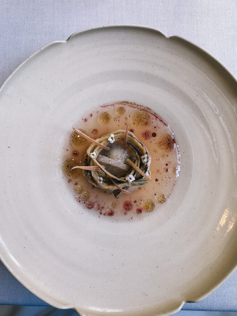 A delicately plated course at Zén featuring a rolled ingredient bundle at the centre of a wide scallop-edged ceramic bowl, surrounded by a light broth with scattered drops of oil and small herb flowers.
