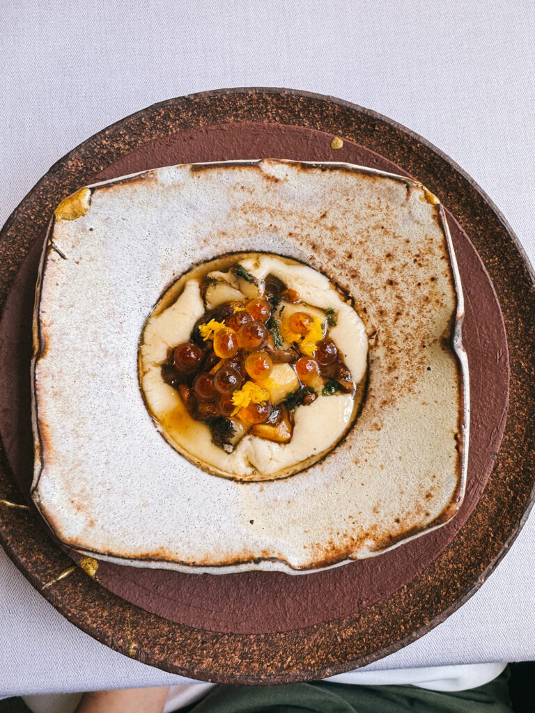 A main course at Zén served on a rustic two-toned ceramic plate, featuring pearls of roe and mushrooms over a cream base, garnished with yellow flower petals.