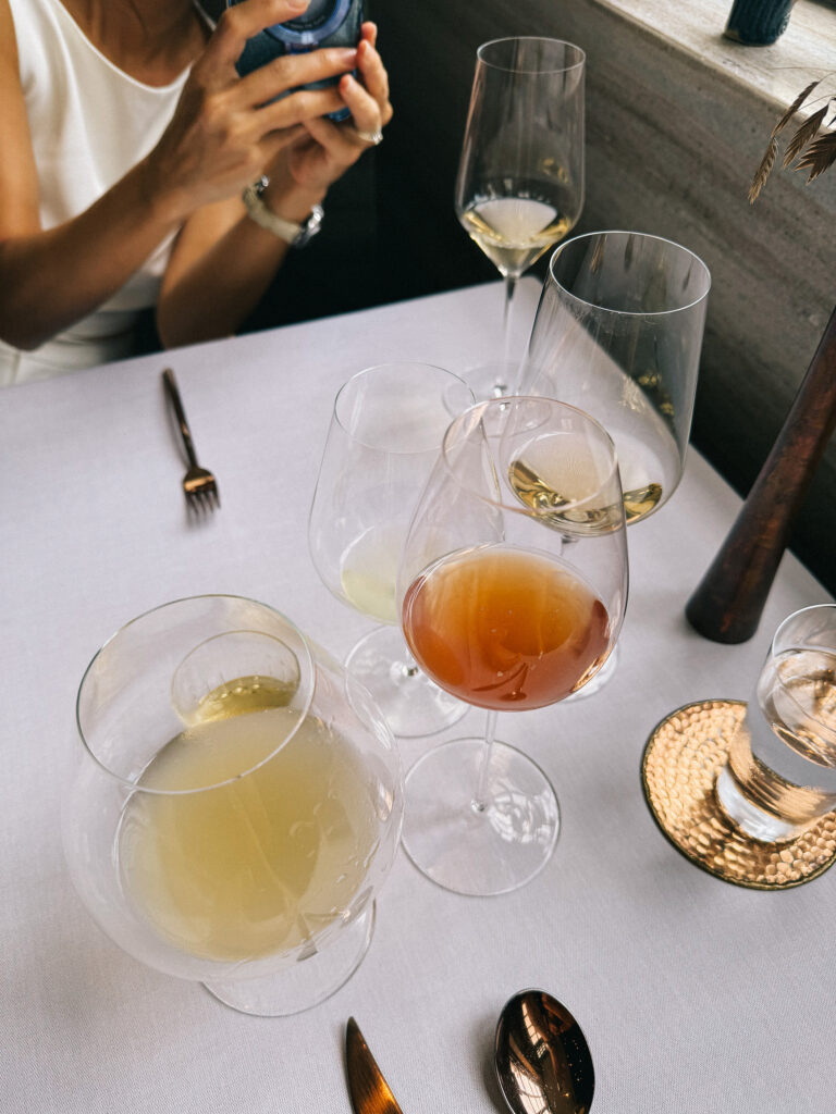 A table setting at Zén's dining room showing the mixed beverage pairing: a pale fermented drink in a tumbler, an amber-toned ferment in a wine glass, and two white wines, alongside gold cutlery on a white linen tablecloth.
