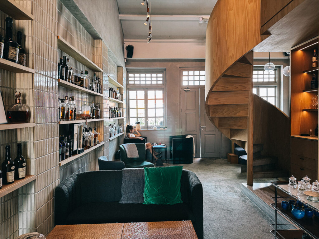 The Living Room on the third floor of Zén, featuring dark velvet sofas, shelves lined with spirit bottles, a spiral wooden staircase and natural light streaming through heritage shophouse windows.
