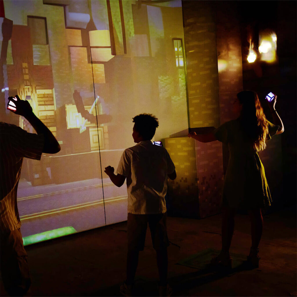 Participants interacting with immersive Minecraft-style digital environment using handheld devices in a dark exhibition space