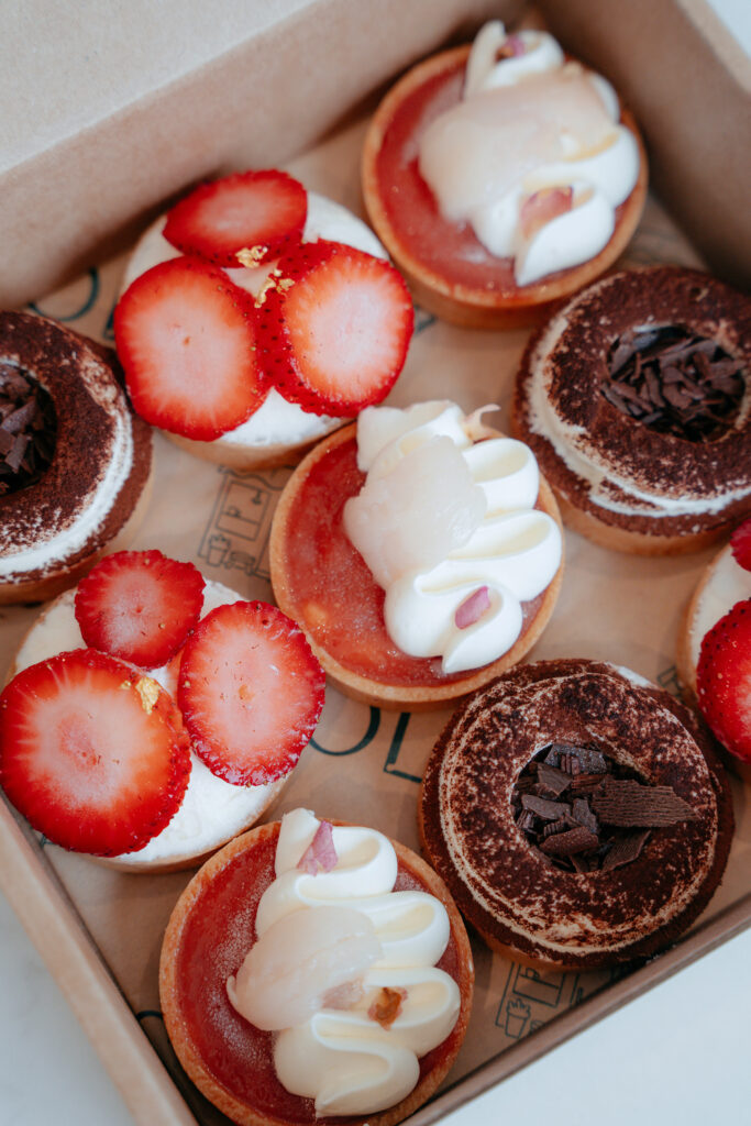 
A box of nine mini tarts from Lola's Cafe featuring Strawberry Pistachio, Pink Guava Lychee and Tiramisu flavours, presented in a branded kraft box