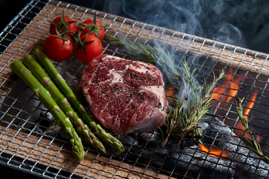 
A seasoned steak cooking over open charcoal flames alongside asparagus, cherry tomatoes and rosemary at Verandah Rooftop Rotisserie