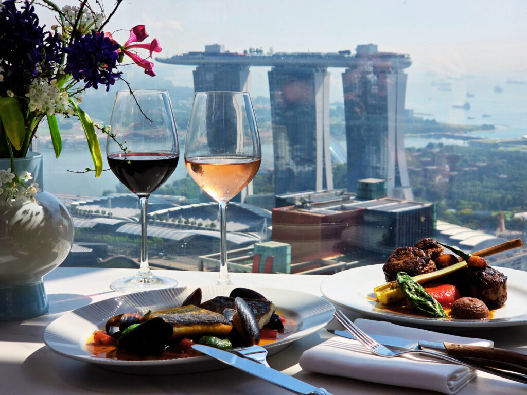 Two plates of Southern European mains and wine glasses set against a panoramic view of Marina Bay Sands from Artemis Grill & Sky Bar, Singapore