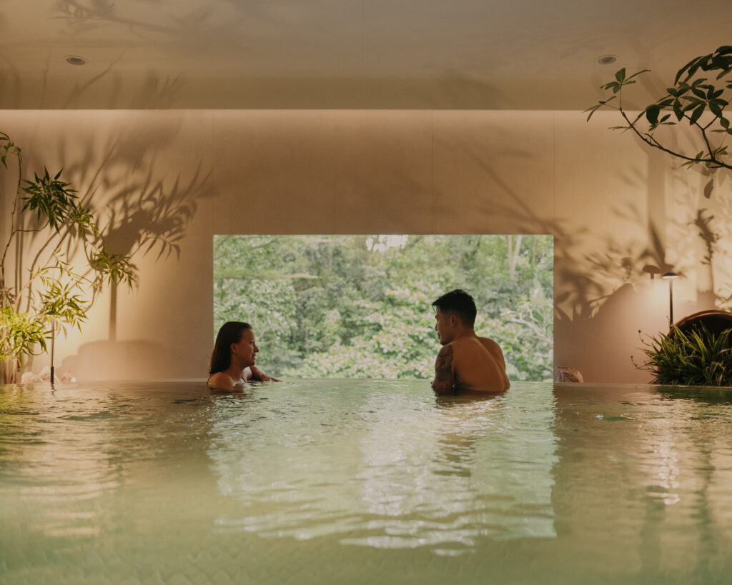 Two guests soaking in the warm pool at Nowhere Baths, Dempsey Hill, with a view of surrounding greenery