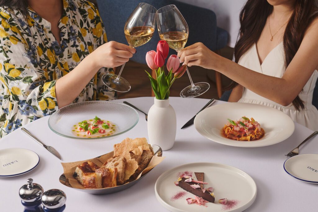 Two diners clinking white wine glasses over a table set with crudo, pasta, chocolate tart and bread at L'Amo Bistrò del Mare, Singapore