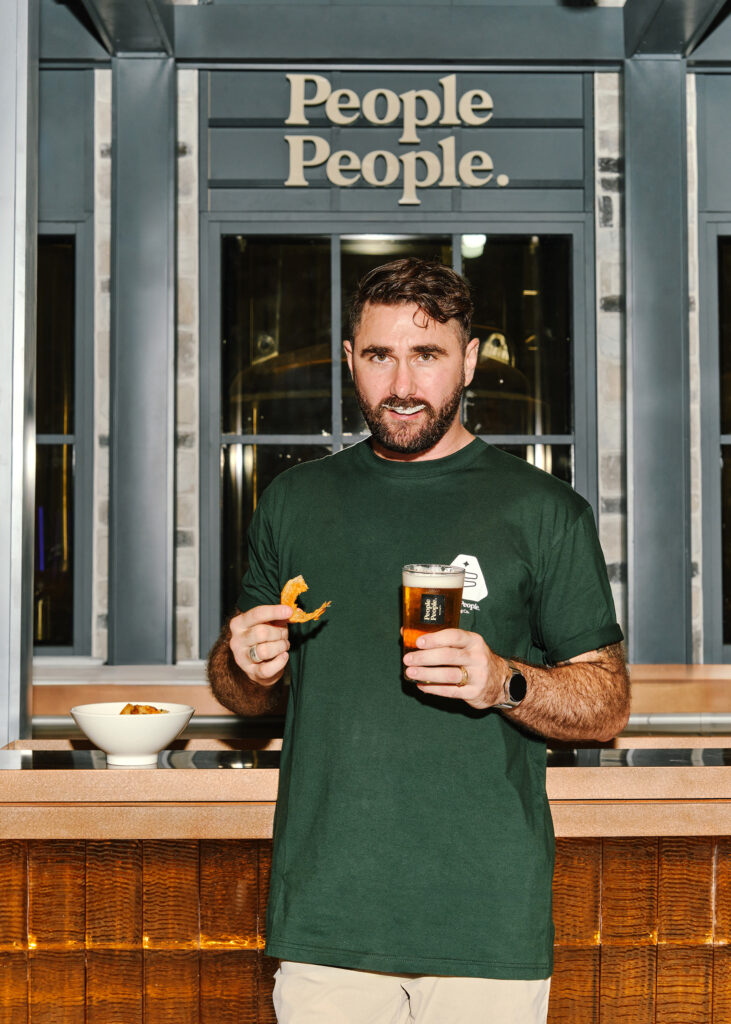 A man in a green People People Brewing Co. t-shirt holds a pint of beer and a piece of fried food at the brewery bar, with copper brewing tanks visible behind him.