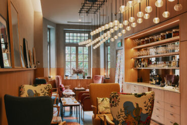 The Kitchen on the first floor of Zén, a warmly lit lounge space with eclectic armchairs in floral and bouclé upholstery, pendant globe lights overhead, and a service bar lined with glassware and fermentation jars along the right wall.