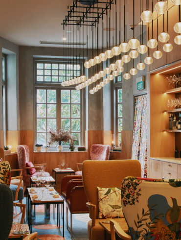 The Kitchen on the first floor of Zén, a warmly lit lounge space with eclectic armchairs in floral and bouclé upholstery, pendant globe lights overhead, and a service bar lined with glassware and fermentation jars along the right wall.