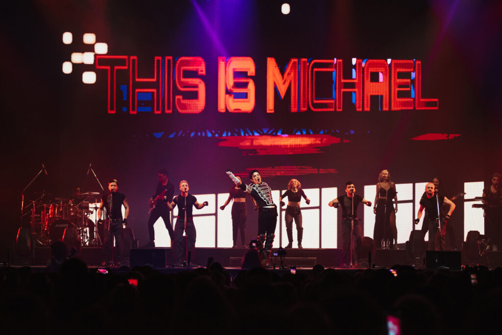 Full stage performance of This Is Michael with dancers and live band, illuminated by bold red visuals and large LED backdrop.
