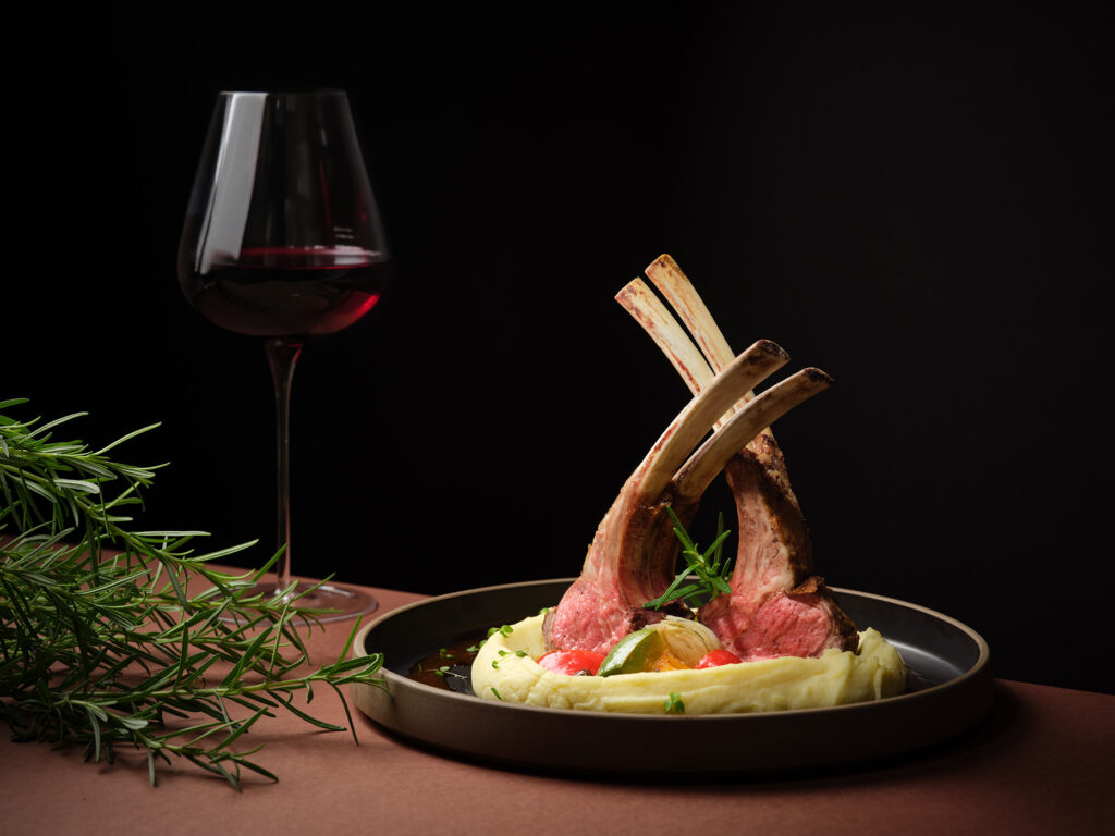 A plated rack of lamb served on creamy mashed potato with natural jus and rosemary garnish, alongside a glass of red wine, at The Lobby Lounge, Frasers House Singapore.