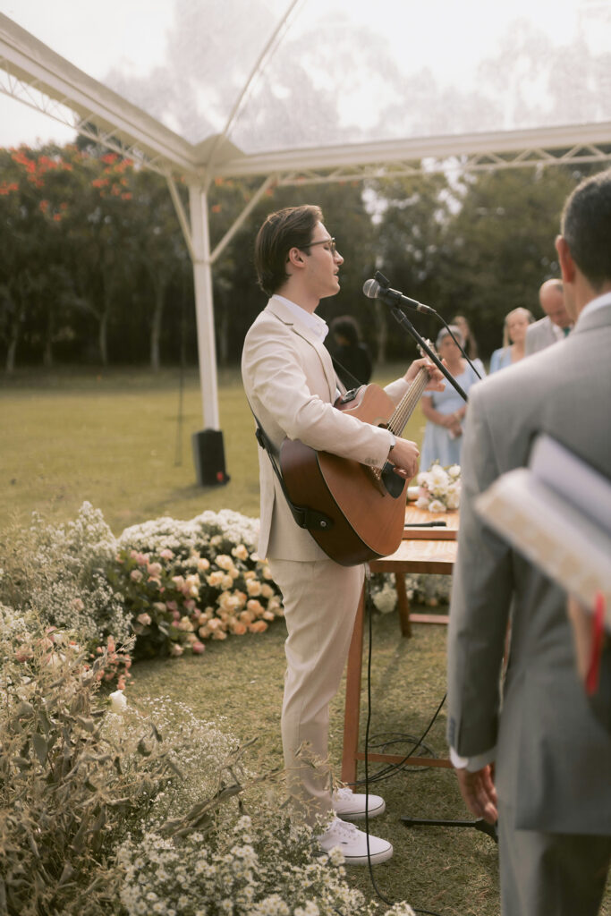 Man in a beige suit playing acoustic guitar and singing at an outdoor wedding ceremony under a canopy, with floral arrangements and guests in the background.