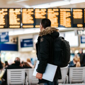 A male traveller holding a laptop looks at a flight information board at an airport terminal.