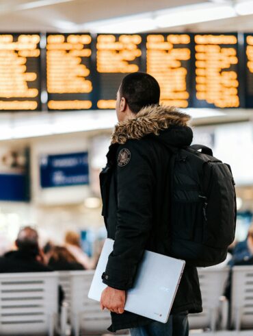 A male traveller holding a laptop looks at a flight information board at an airport terminal.