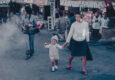 A woman holds a young child's hand while walking through a busy street market, captured in a vintage colour photograph