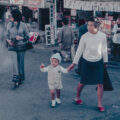 A woman holds a young child's hand while walking through a busy street market, captured in a vintage colour photograph