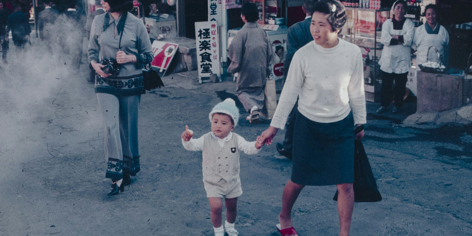A woman holds a young child's hand while walking through a busy street market, captured in a vintage colour photograph