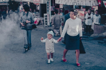 A woman holds a young child's hand while walking through a busy street market, captured in a vintage colour photograph